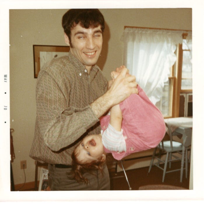 A young man with black hair swings a toddler wearing pink upside down. The photo is marked May 1970. A young man with black hair swings a toddler wearing pink upside down. The photo is marked May 1970.