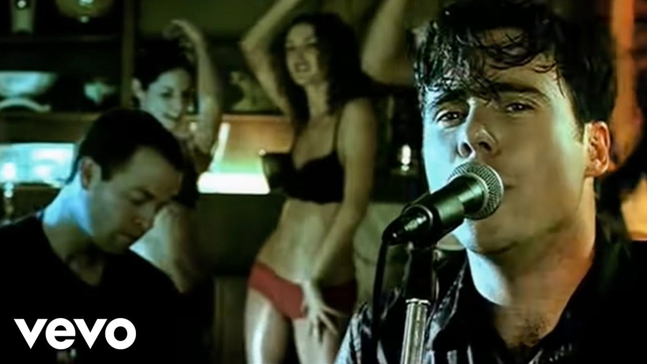 White man sings into a microphone wearing a black shirt and sweating, another white man is playing drums and several white women in underwear dance in the background.