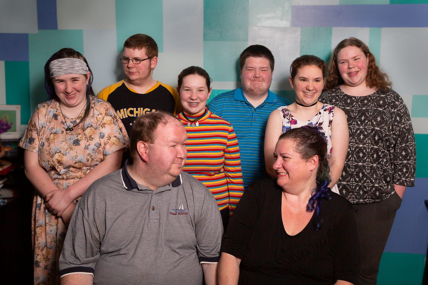 Family portrait featuring a man and a woman in their 40’s sitting and looking at each other and smiling. Behind them stand their 6 children ages 13-20. The children are all mid laugh. The family is all white skinned with brown hair. 