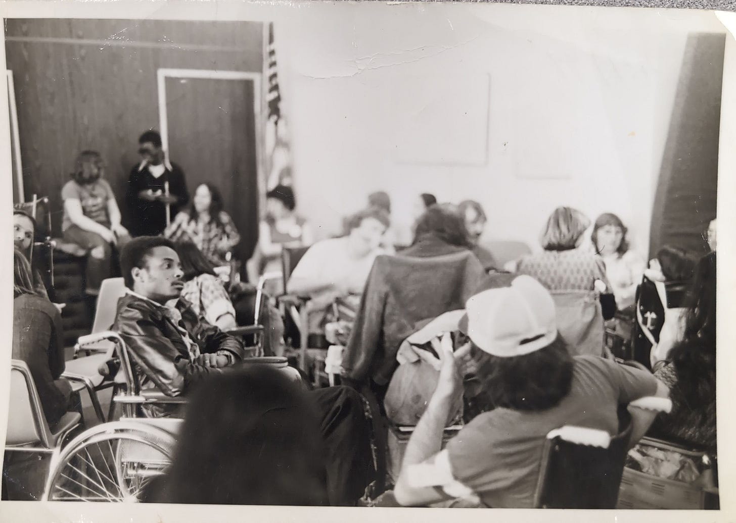A black and white image from the federal sit in, featuring a lot of disabled people packed into a room, Brad Lomax a Black man in his wheelchair to the right of the image, his arms folded as he stares at someone across the room