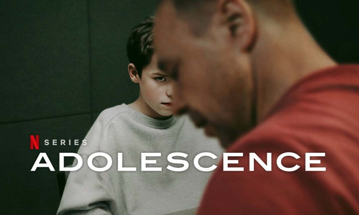 Promotional image for the Netflix series "Adolescence," showing a boy with dark hair sitting at a table, seen past the out-of-focus shoulder and head of a man in a red shirt in the foreground. The Netflix "N Series" logo and the title "ADOLESCENCE" appear in white text at the bottom left.