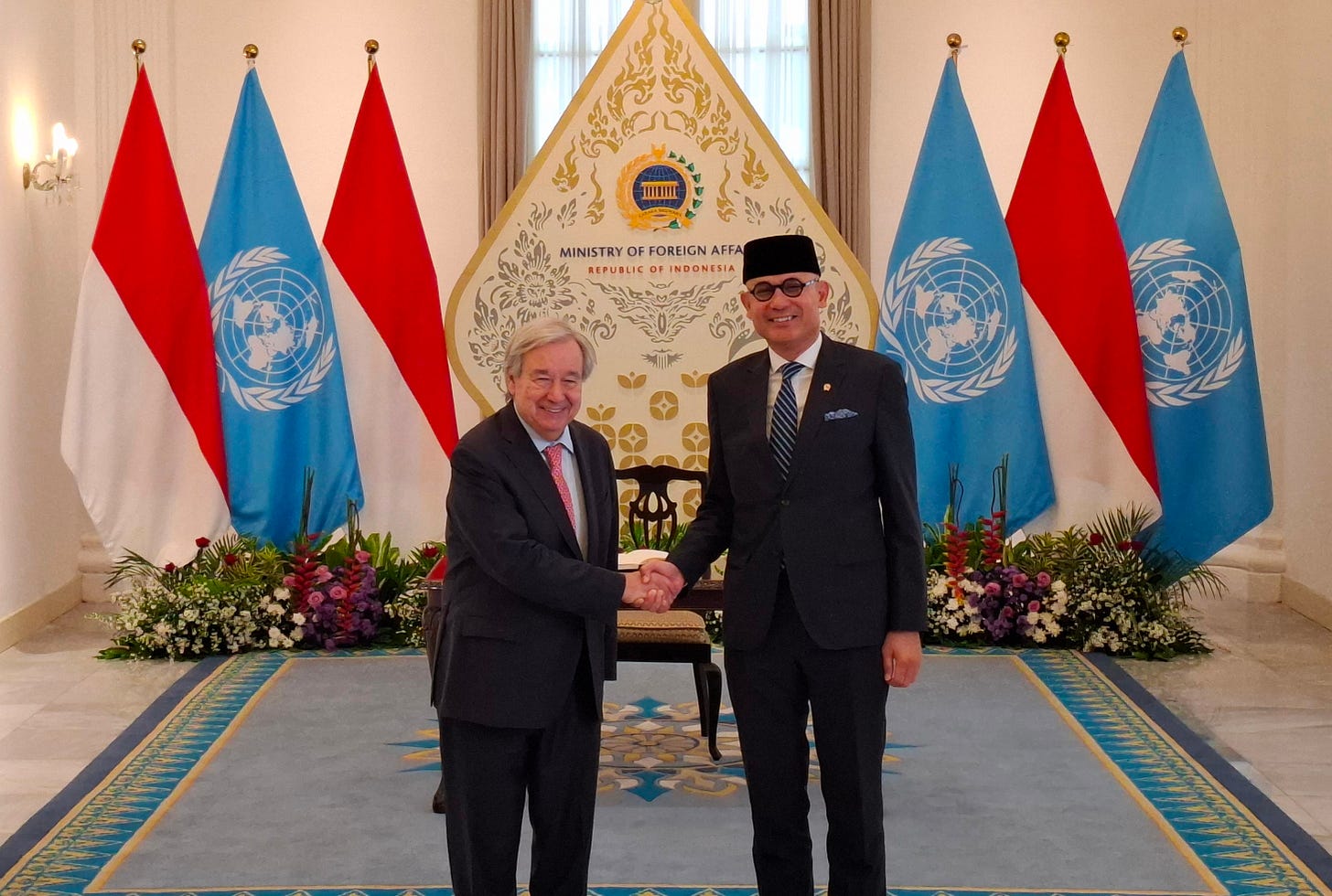 Two people stand in front of the UN and Indonesian flags, shaking hands. On the left is UN Secretary-General António Guterres, and on the right is Indonesian Foreign Affairs Minister Arrmanatha Nasir. Minister Nasir welcomes Guterres to the Pancasila Building in Jakarta on Monday (19 October) for foreign policy talks with diplomats and attendees at the Foreign Affairs Ministry.