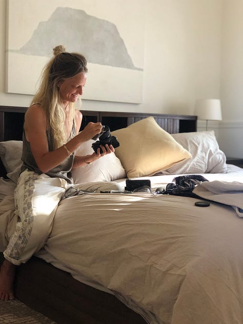 One row of three images, left to right: Woman sitting at dining table presenting her laptop screen showing her creative designs for stationery with bubbles of light on the computer coming through the window in the background; Photo of woman’s back as she holds her digital camera with one hand up to her shoulder, looking off into the desert mountain landscape from atop a trail; Photo of woman cleaning her camera lenses sitting on a bed with sunlight streaming in through the window and an abstract painting hanging on the wall behind her.