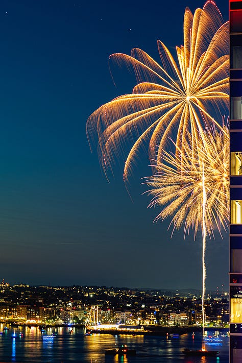 Night fireworks exploding beside Seaport skyscraper with Boston skyline and harbor reflections