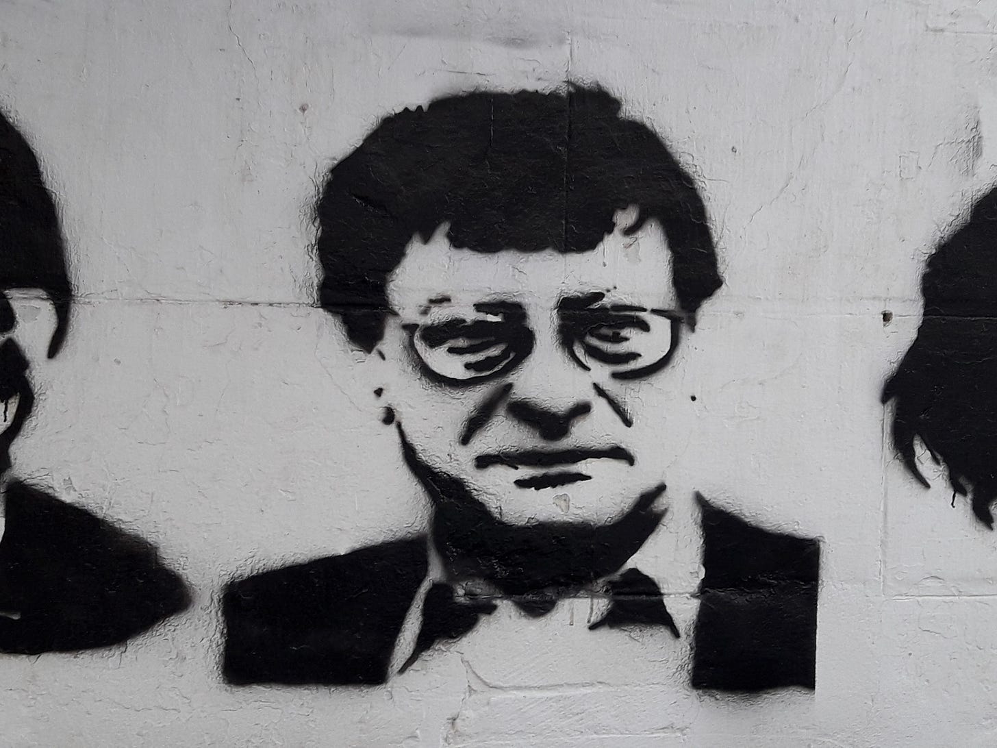 Black stencil graffiti portrait of a man wearing glasses on a white wall, with parts of two other faces partially visible on either side.