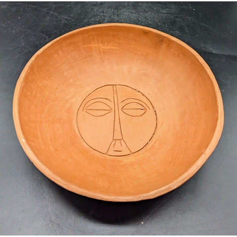 May include: A terracotta bowl with a carved minimalist face design. The bowl is round and shallow, with a warm, earthy tone. The face is centrally located and features simple lines for the eyes, nose, and mouth.  The bowl's surface shows subtle variations in texture and color, characteristic of handmade pottery. This decorative bowl is perfect for adding a touch of rustic elegance to any space.