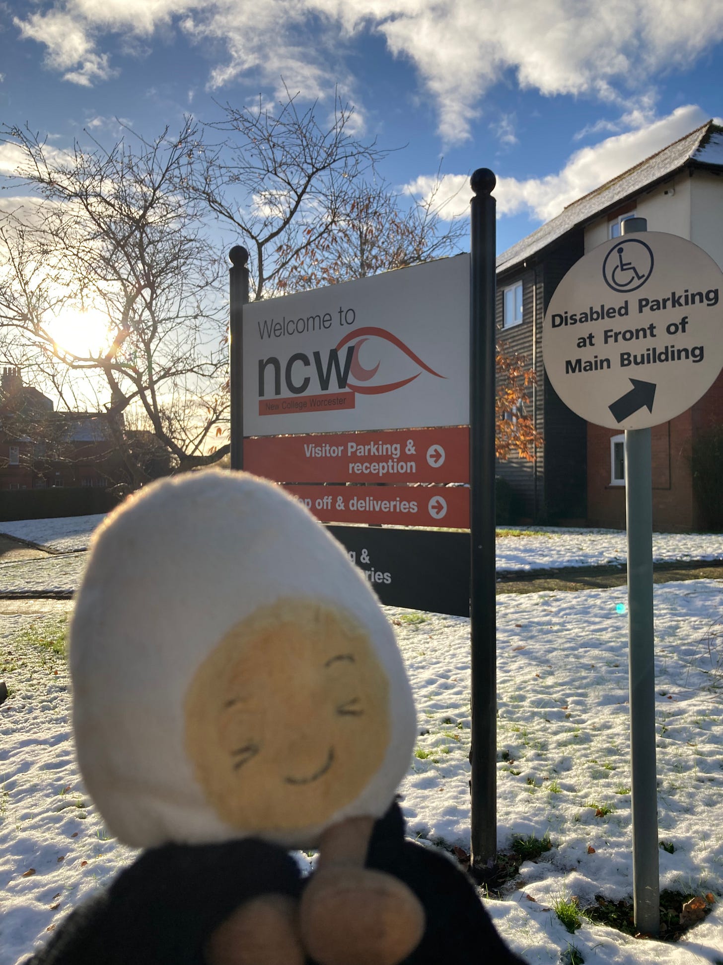 Dippy the soft toy egg in front of a sign saying welcome to ncw new College Worcester, in a carpark. The ground has a light covering of snow and the sun is shining brightly through the empty trees.