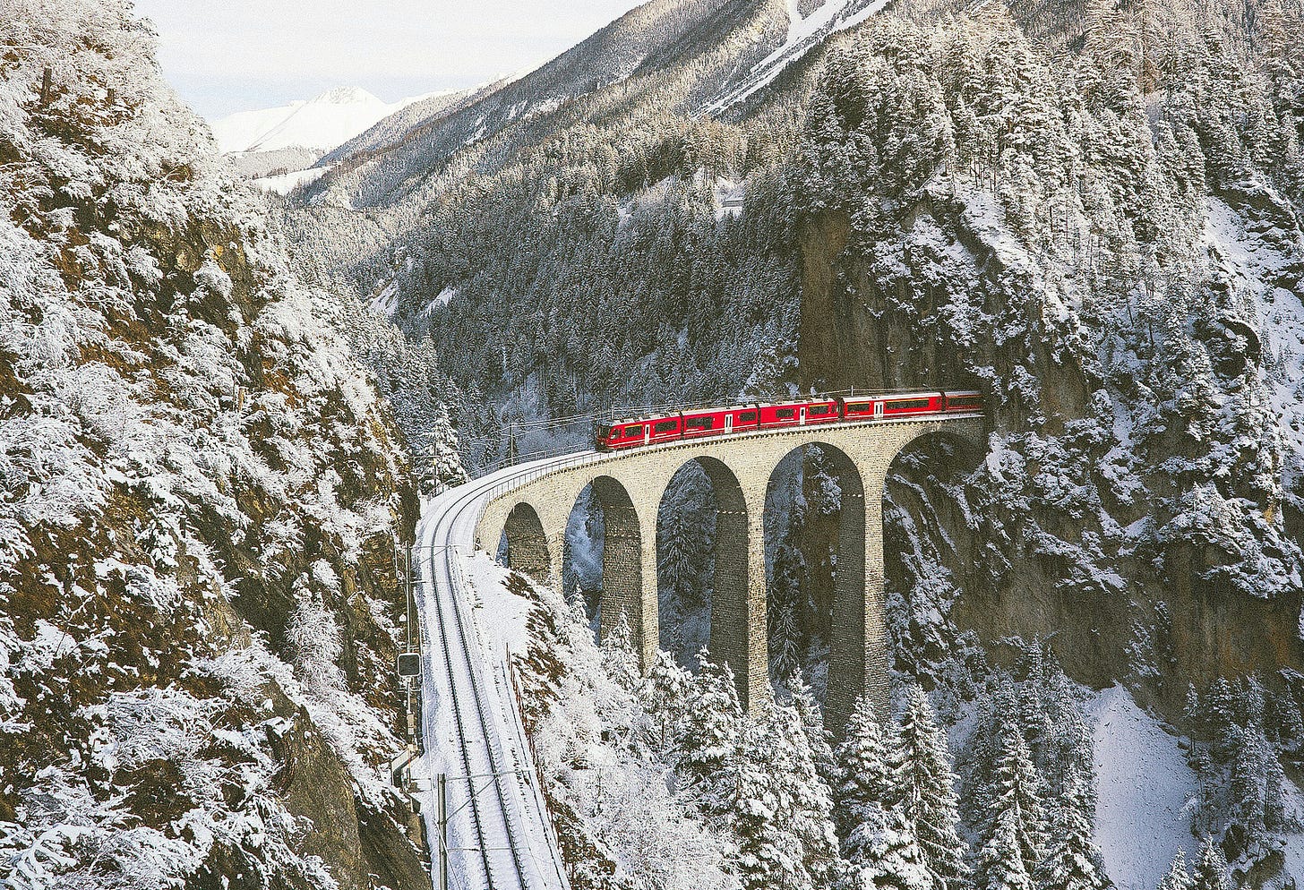 A bright red train exiting a mountainside tunnel onto a high brick bridge over a crevasse, set against a bright but brown, white and dark green, snow-covered mountain valley landscape, and moving along snow-covered tracks that turn after the bridge and continue through the bottom of the image. A bright red train exiting a mountainside tunnel onto a high brick bridge over a crevasse, set against a bright but brown, white and dark green, snow-covered mountain valley landscape, and moving along snow-covered tracks that turn after the bridge and continue through the bottom of the image.