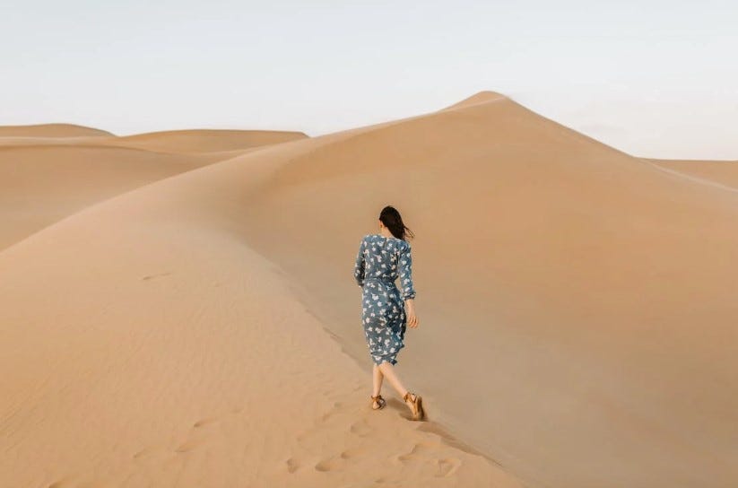 A lone woman wearing a blue floral dress and sandals walks along the crest of a sand dune in a vast desert. The image shows her from behind as she faces the horizon, with more sand dunes visible in the distance under a pale sky.