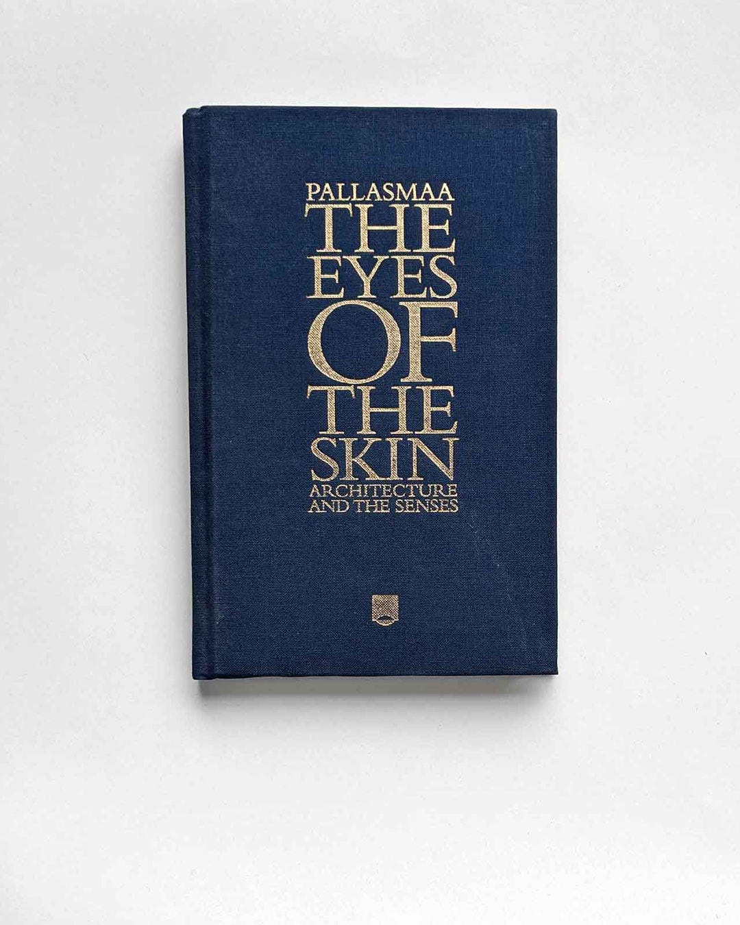 A hardcover book photographed flat against a white surface. The cover is deep navy cloth with gold foil lettering reading: "PALLASMAA / THE EYES OF THE SKIN / ARCHITECTURE AND THE SENSES," with a small publisher's shield emblem at the bottom center.