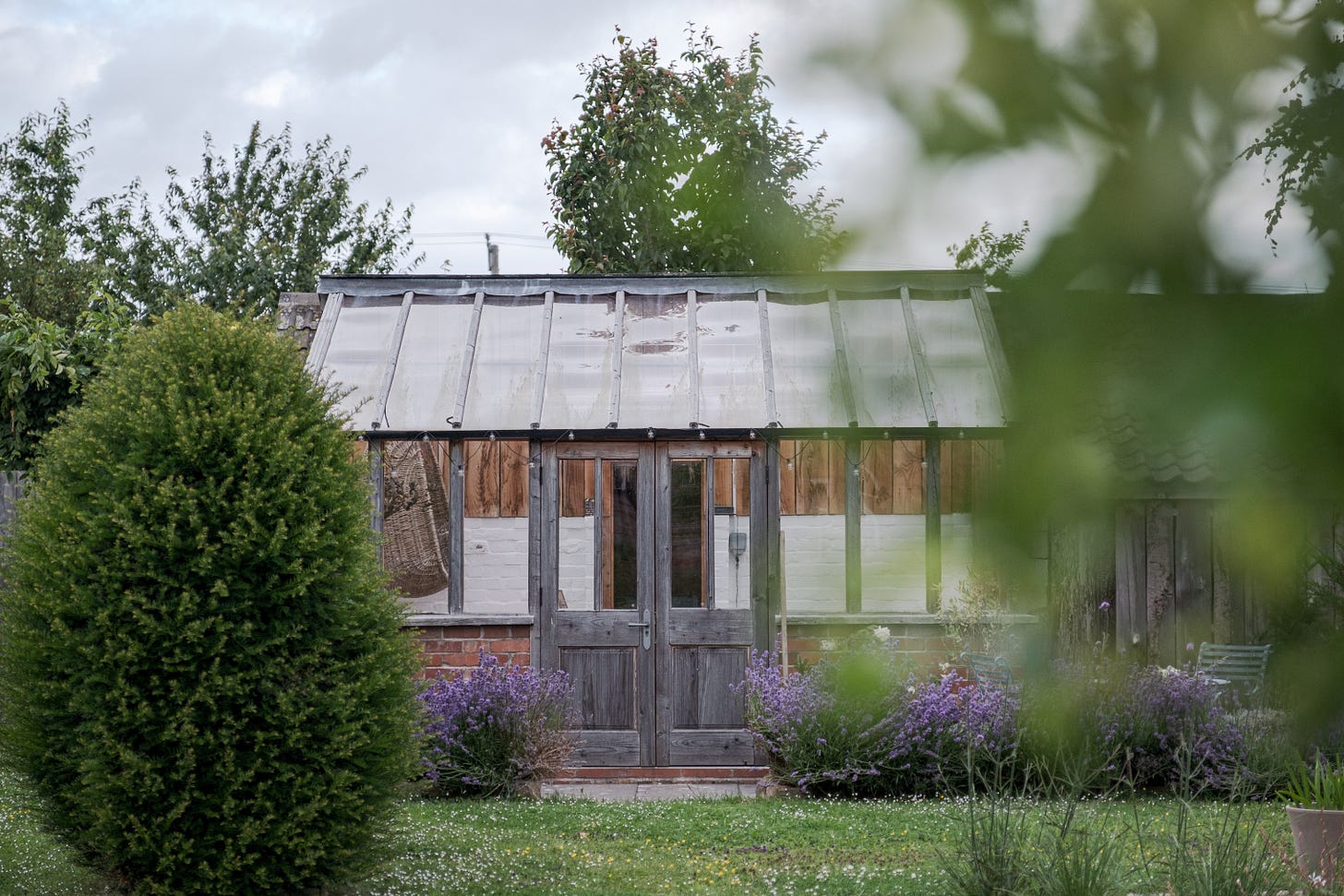 Photo of wooden garden room with painted white brick walls inside and hanging basket chair, in front of the room are bushy yew hedges and flowering lavender