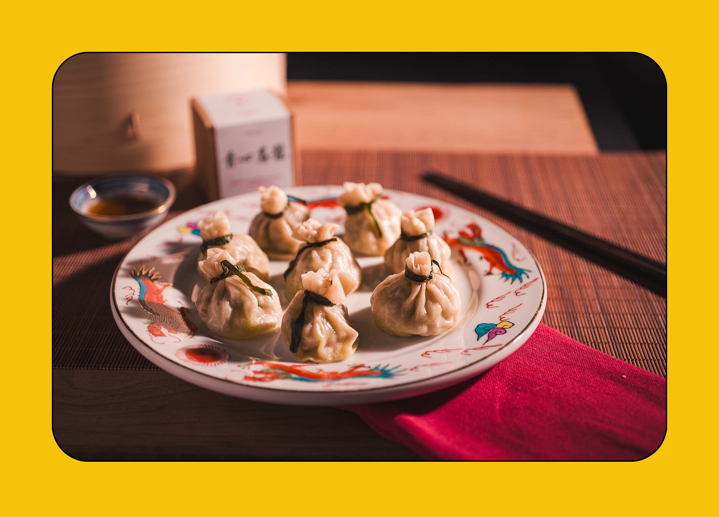 A plate of steamed dumplings is arranged on a decorative dish with a red napkin, soy sauce, and a bamboo steamer in the background.