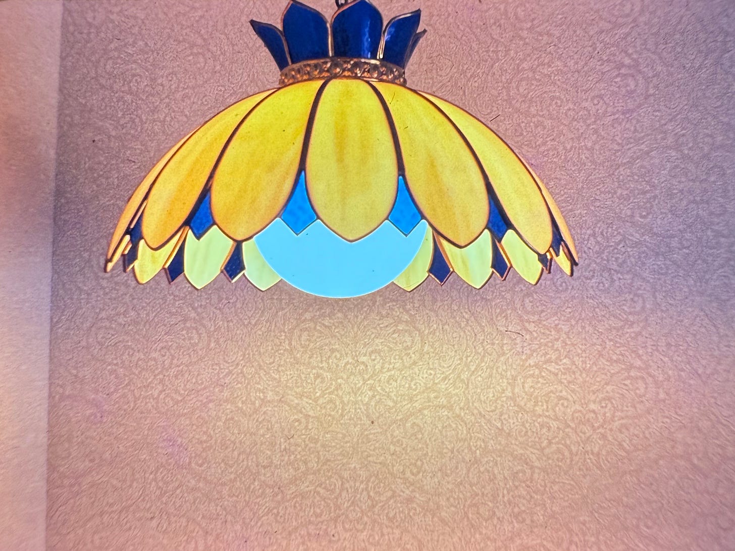 a blue and yellow slag glass lamp in the shape of petals of a flower that hang down over a large, spherical, robin's-egg-blue bulb. Behind the lamp is intricately designed pale wallpaper. a blue and yellow slag glass lamp in the shape of petals of a flower that hang down over a large, spherical, robin's-egg-blue bulb. Behind the lamp is intricately designed pale wallpaper.