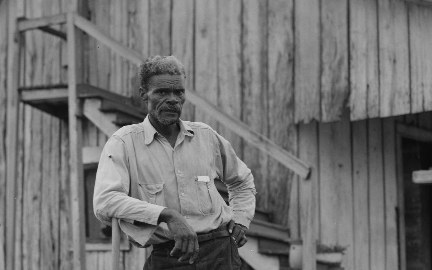 Black and white photograph shows a black man with a beard leaning on an axe, wooden house behind him Black and white photograph shows a black man with a beard leaning on an axe, wooden house behind him