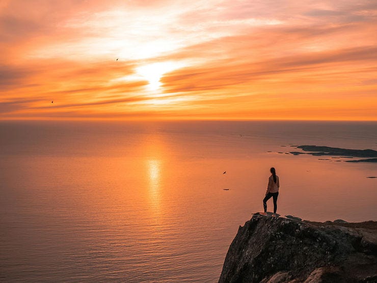 Woman standing on a cliff overlooking an orange and pink sunset on the ocean. Woman standing on a cliff overlooking an orange and pink sunset on the ocean.