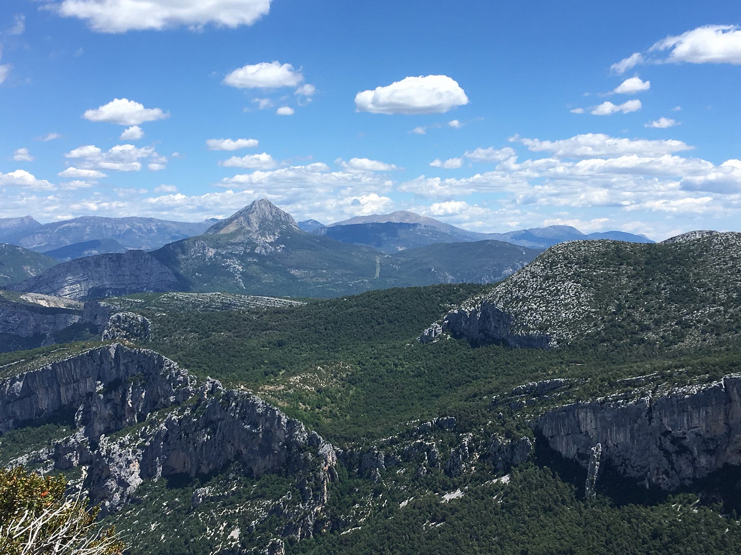A view of a mountain range stretching into the distance. Above it, the sky is blue and dotted with clouds.