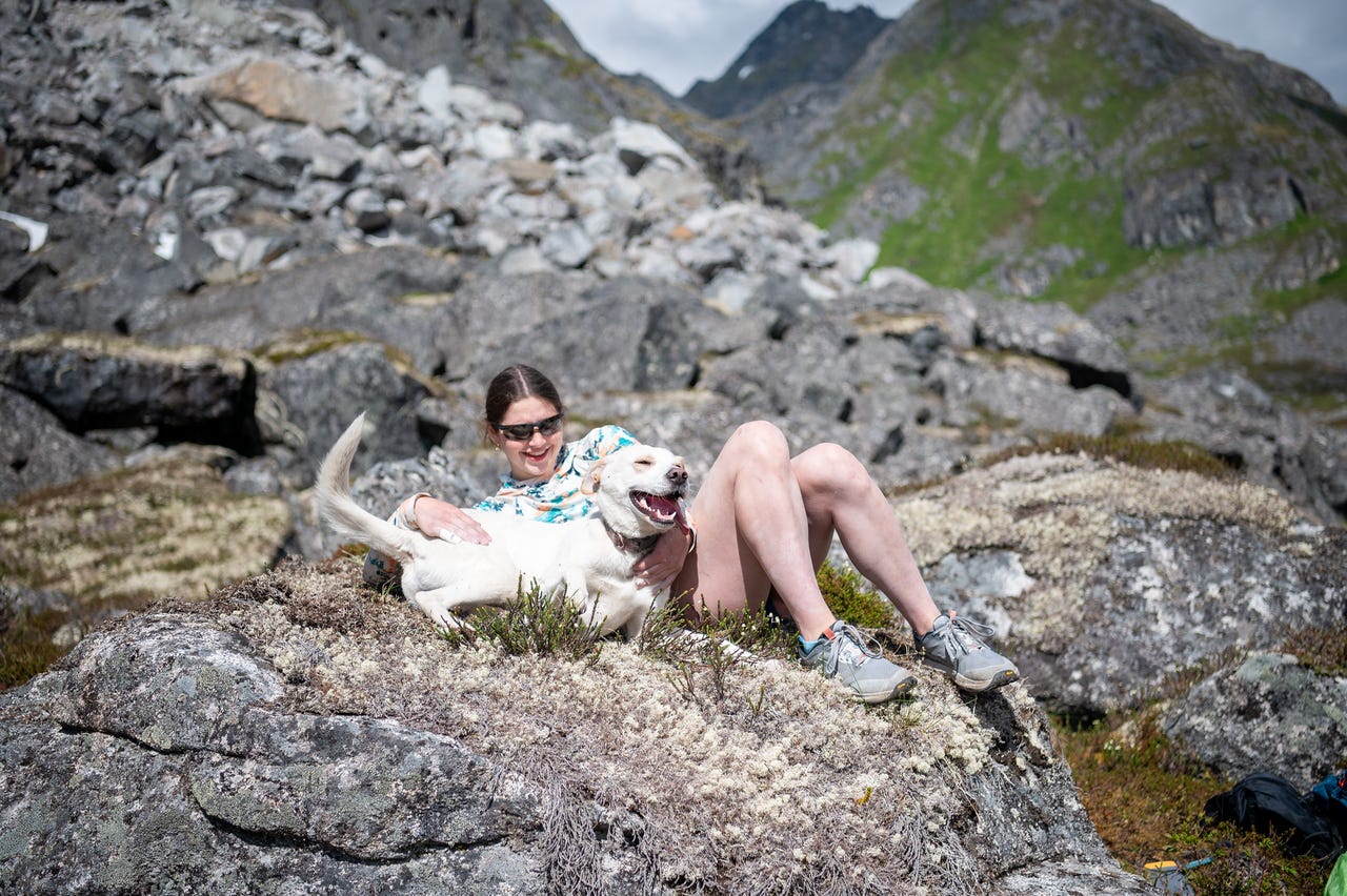 A girl in sunglasses and a colorful shirt sits on a lichen-covered rock with her knees up, laughing as she wraps an arm around a white dog lying across her lap, the dog's mouth open in a wide pant that reads as a grin, tail mid-wag. Behind them, grey talus and a green-sloped peak rise under an overcast-bright sky. A girl in sunglasses and a colorful shirt sits on a lichen-covered rock with her knees up, laughing as she wraps an arm around a white dog lying across her lap, the dog's mouth open in a wide pant that reads as a grin, tail mid-wag. Behind them, grey talus and a green-sloped peak rise under an overcast-bright sky.
