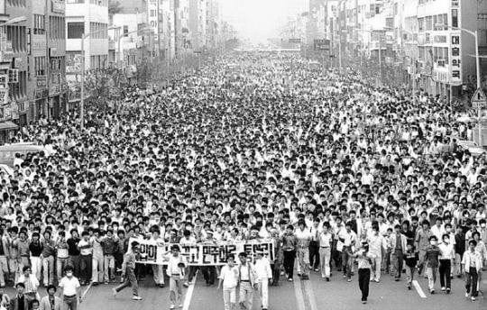 Black-and-white photo of an enormous crowd of people filling a wide city street, marching shoulder to shoulder between rows of buildings; at the front, demonstrators hold a large Korean banner as they protest during the 1980 Gwangju Uprising. Black-and-white photo of an enormous crowd of people filling a wide city street, marching shoulder to shoulder between rows of buildings; at the front, demonstrators hold a large Korean banner as they protest during the 1980 Gwangju Uprising.