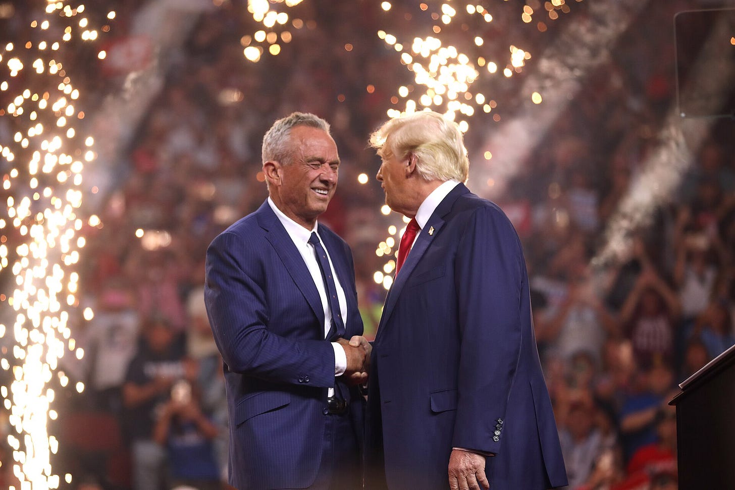 RFK shaking hands with Trump, both in suits, in front of lights
