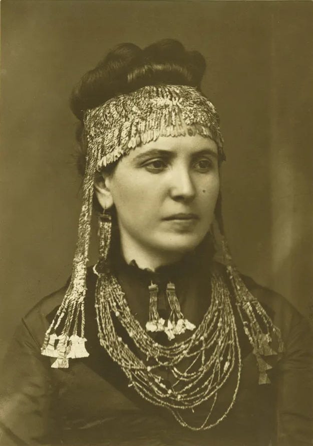 A woman poses for a portrait wearing traditional jewelry, including a headpiece with metallic fringe and layered beaded necklaces.