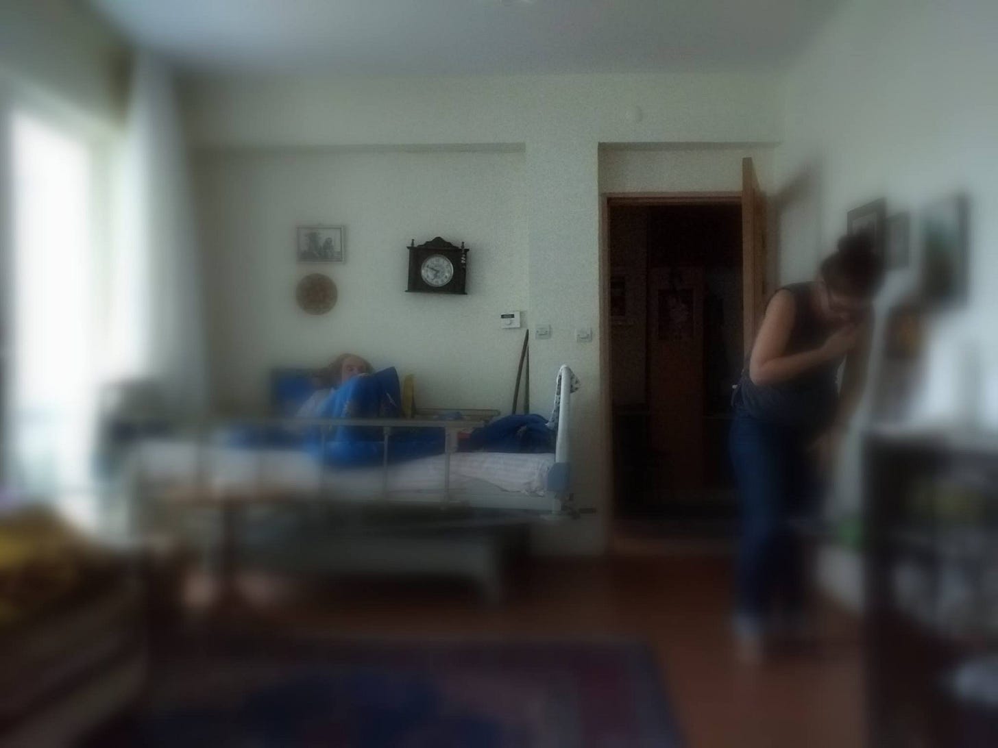 Biraz flu bir fotoğraf. Bir evin odası, karşıda bir duvarın dibinde bir hasta yatağı, yatakta kaykılmış oturan bir kadın. Duvarda bir saat, bir siyah-beyaz fotoğraf. Sağda telefonda konuşan bir genç kadın. 