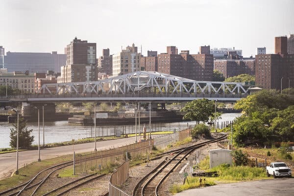 A silver bridge spans the Harlem River, with train tracks in the foreground and many brown and beige buildings on the other side of the river. A silver bridge spans the Harlem River, with train tracks in the foreground and many brown and beige buildings on the other side of the river.