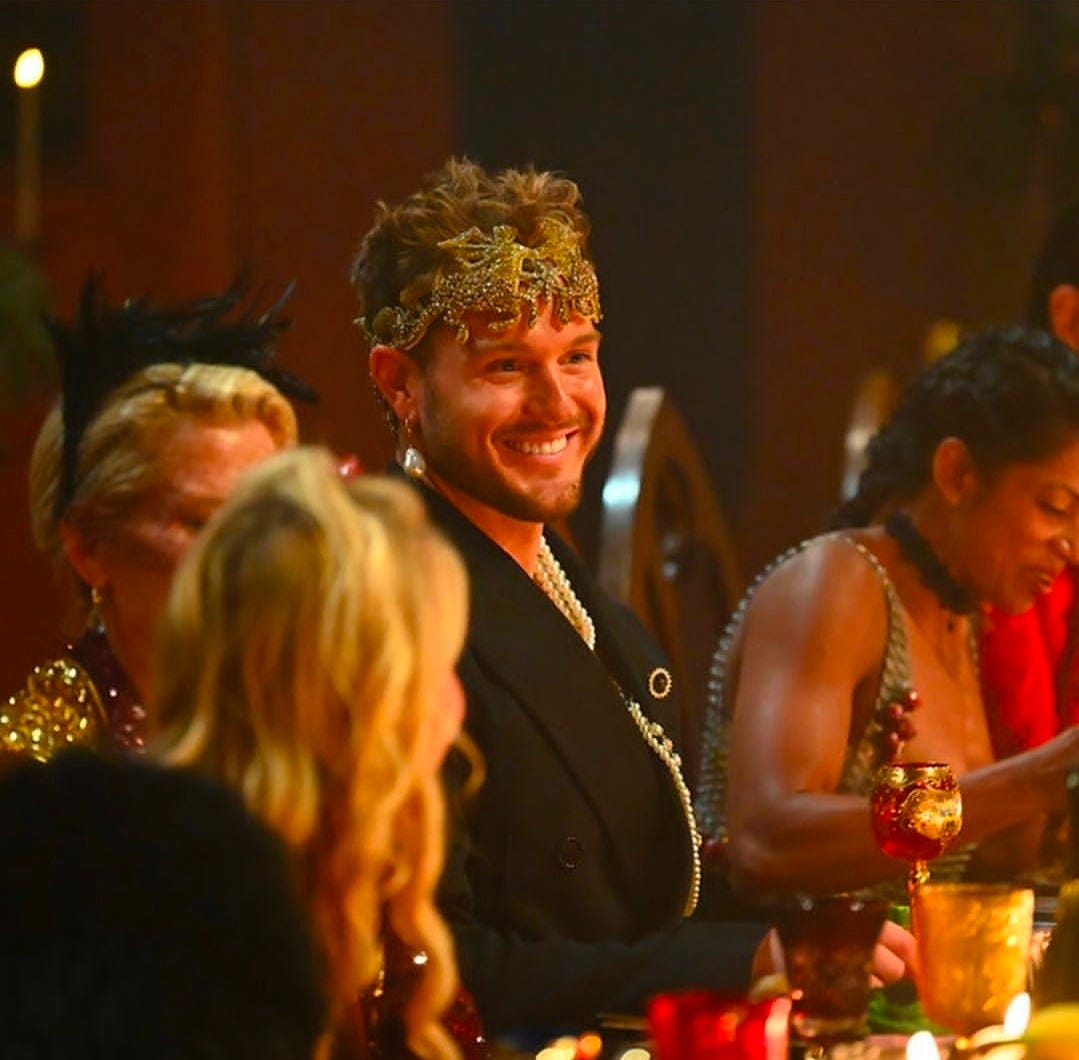 Close-up of Colton at the banquet table smiling, crown visible, pearl harness draped over bare chest under open blazer.