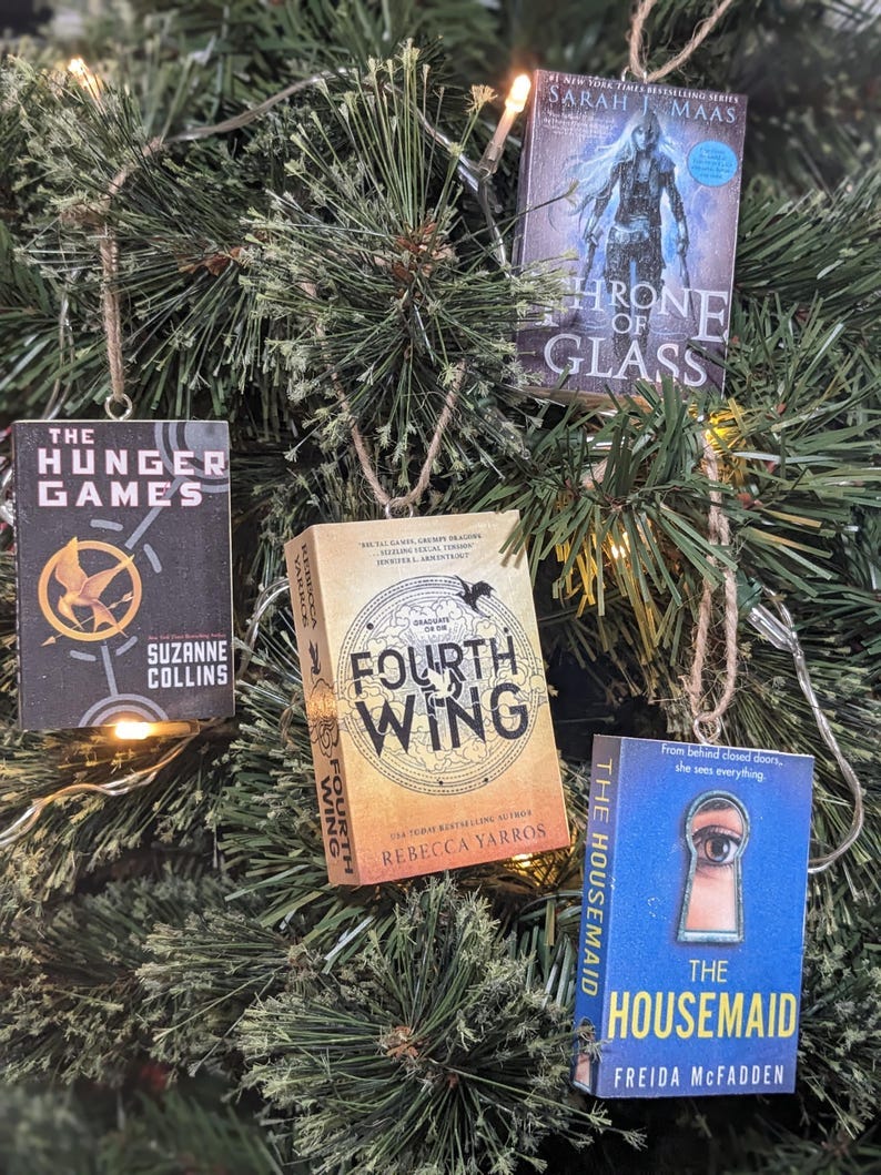 May include: Four book ornaments hanging on a green Christmas tree. The ornaments are made from book covers and include 'The Hunger Games' by Suzanne Collins, 'Throne of Glass' by Sarah J. Maas, 'Fourth Wing' by Rebecca Yarros, and 'The Housemaid' by Freida McFadden.