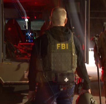FBI agent wearing tactical gear and holding a helmet, standing near an open vehicle door illuminated by red lights. FBI agent wearing tactical gear and holding a helmet, standing near an open vehicle door illuminated by red lights.