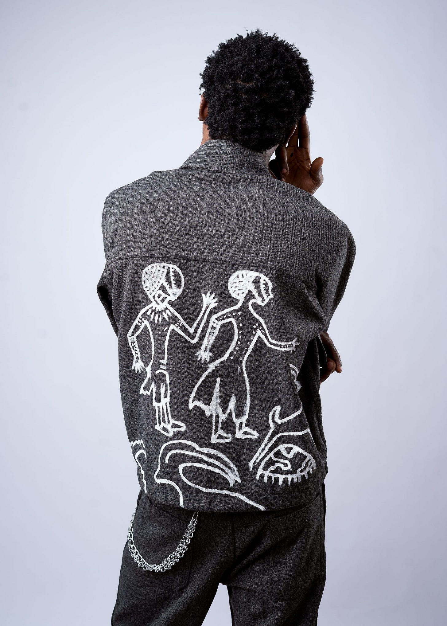 Seen from behind, a model raises a hand beside a charcoal Heritage jacket hand-painted with two white dancing figures Seen from behind, a model raises a hand beside a charcoal Heritage jacket hand-painted with two white dancing figures