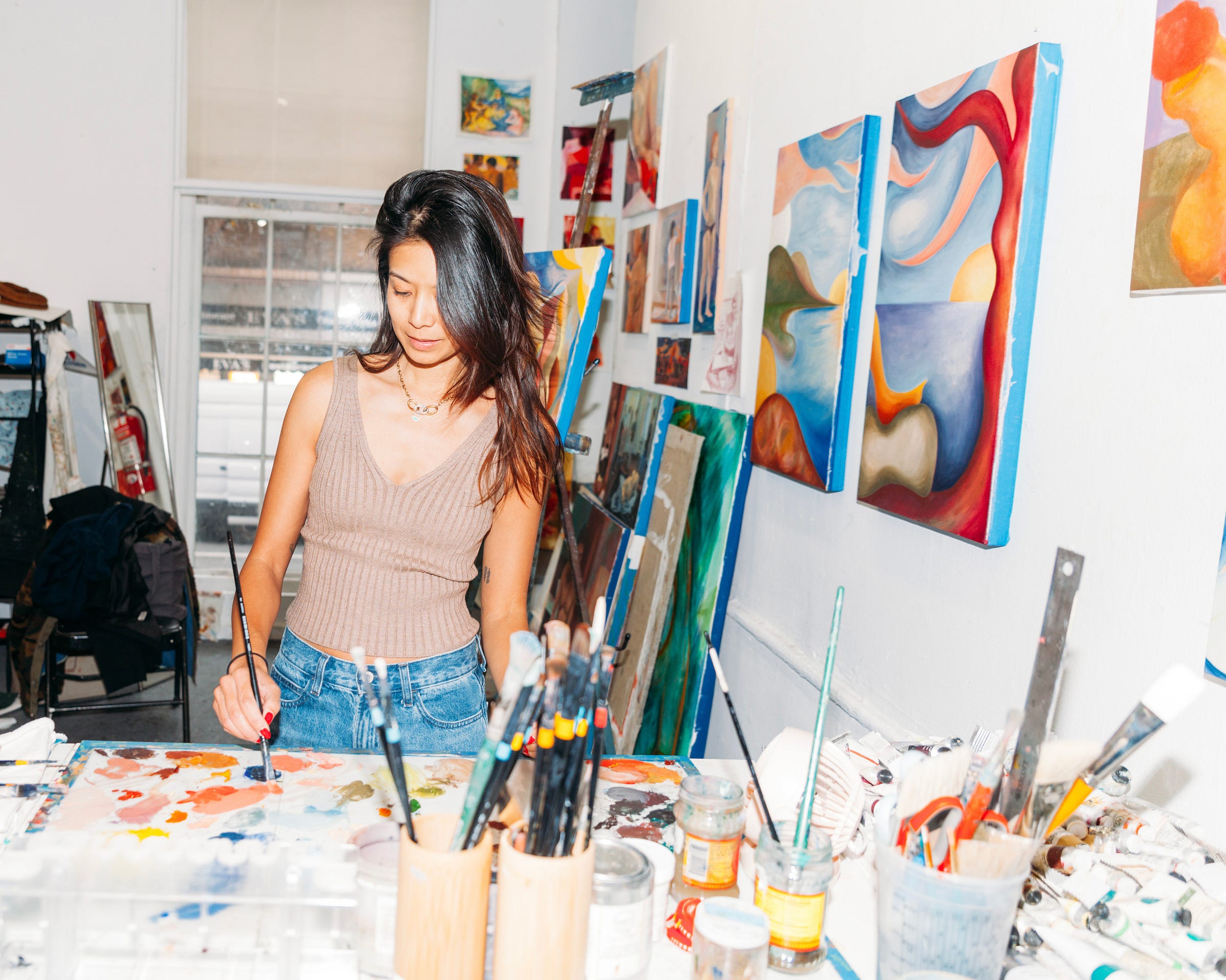 Artist Alex Maceda sits at a paint-covered studio table, working with a brush while colorful abstract paintings and studio materials surround her. Artist Alex Maceda sits at a paint-covered studio table, working with a brush while colorful abstract paintings and studio materials surround her.
