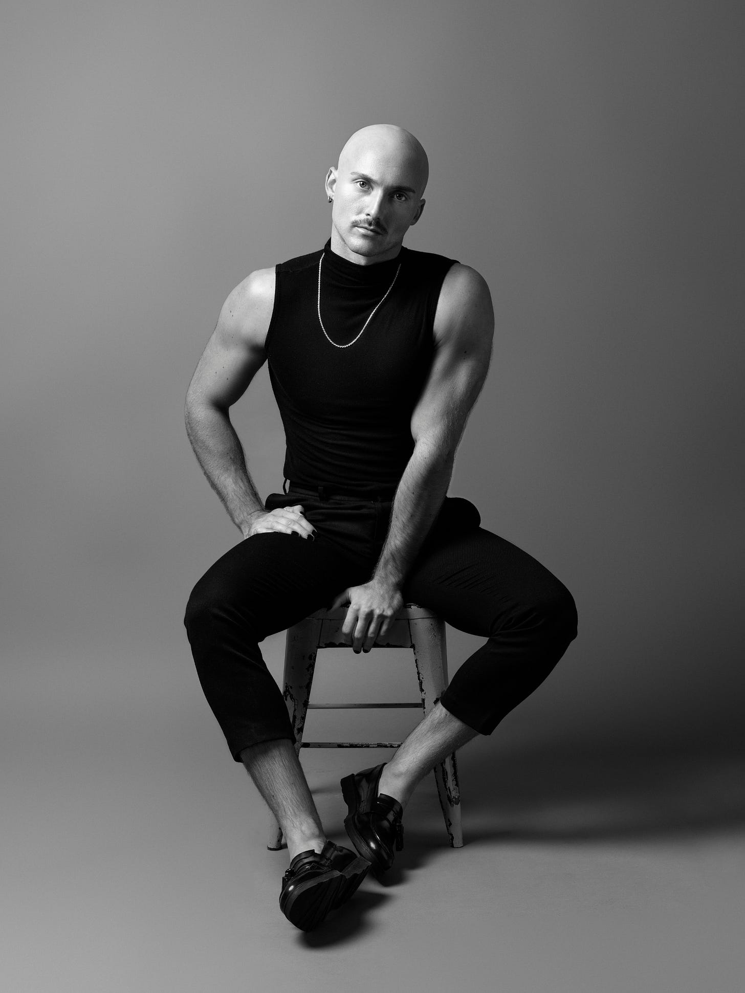 Seated on a stool, Logan is a white person with a mustache and shaved head, wearing a black sleeveless top, silver chain necklace, black pants, and black shoes. They stare directly into the camera without smiling. Photo credit: Hampus Danielsson Seated on a stool, Logan is a white person with a mustache and shaved head, wearing a black sleeveless top, silver chain necklace, black pants, and black shoes. They stare directly into the camera without smiling. Photo credit: Hampus Danielsson