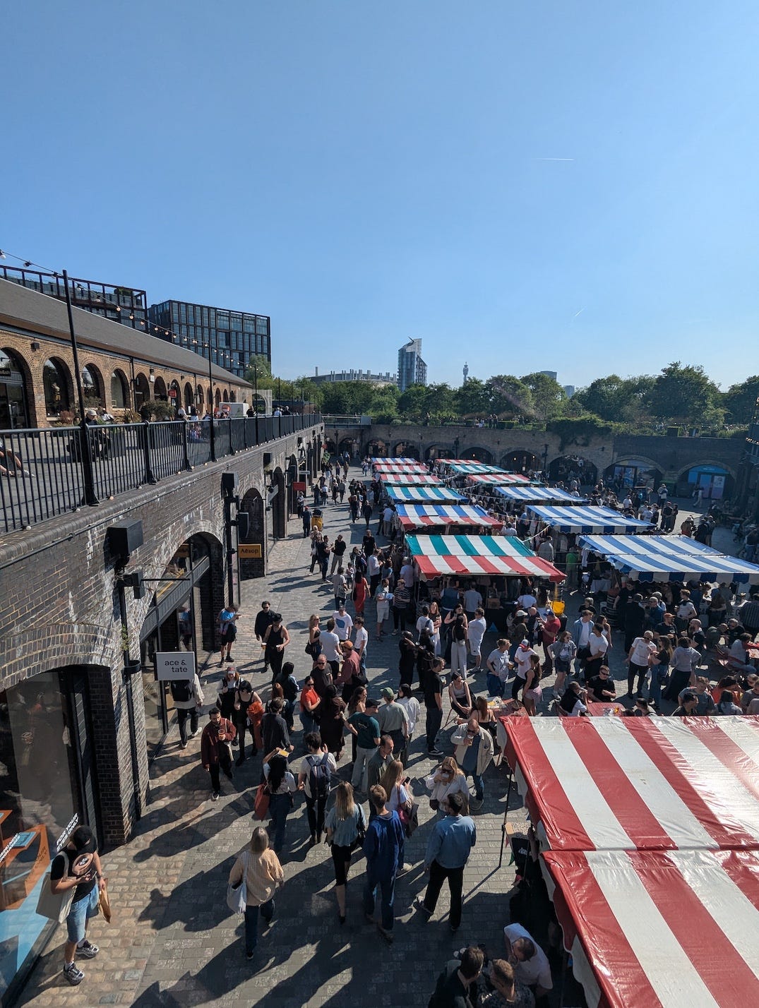 Aerial view of a busy outdoor market at Coal Drops Yard on a sunny day, with colourful striped market awnings and crowds of people milling around the Victorian brick arches.