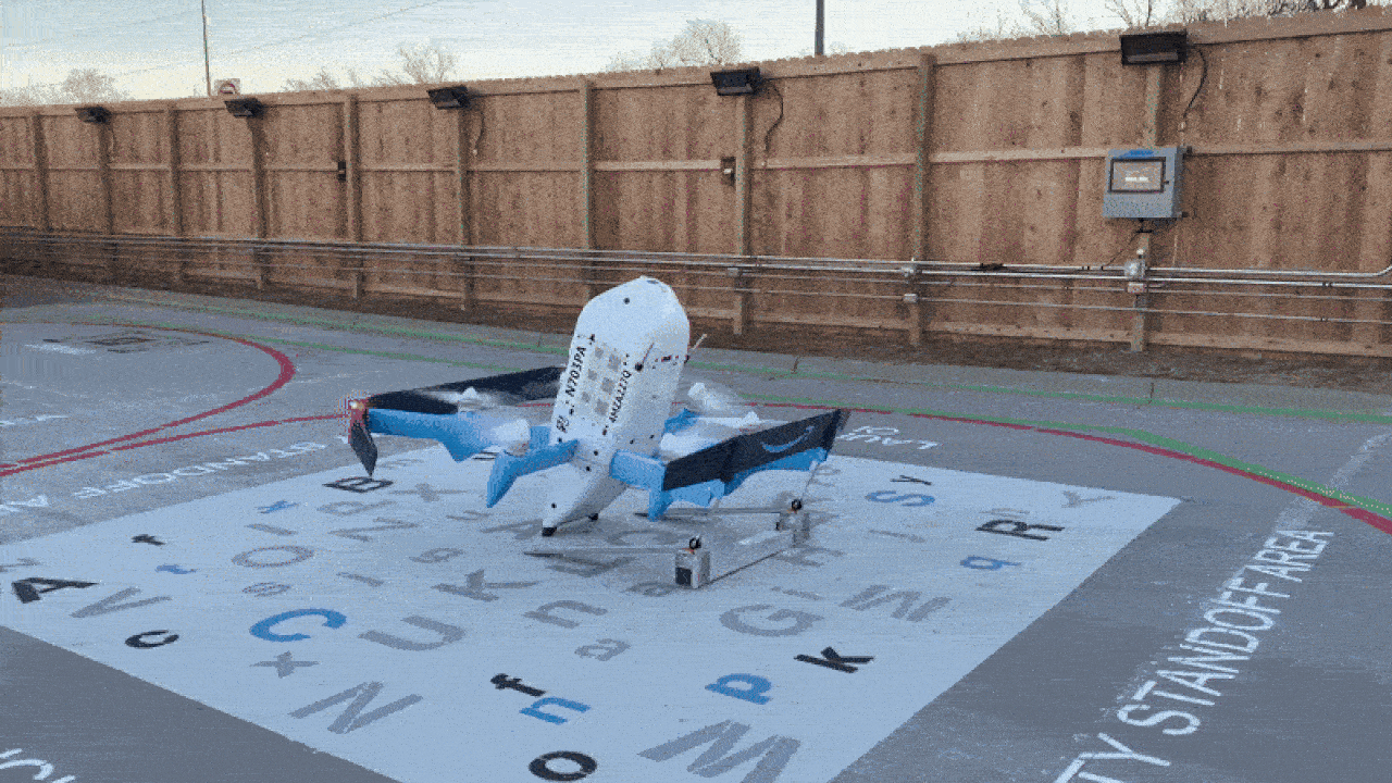 A white and blue drone with multiple propellers stands on a landing pad marked with various letters and labeled "SAFETY STANDOFF AREA" on the right, surrounded by a wooden fence. A white and blue drone with multiple propellers stands on a landing pad marked with various letters and labeled "SAFETY STANDOFF AREA" on the right, surrounded by a wooden fence.