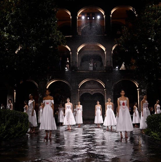 Women standing in stark white dresses in a rain wet courtyard of a dramatic building at night. The archways of the building are underlit, creating illuminated archways
