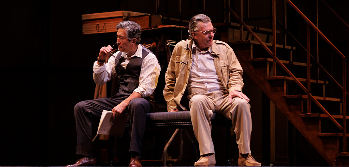 Ying Ruocheng and Arthur Miller sitting together on a bench, facing away from one another. Ying holds a script loosely in one hand, and is taking off his glasses with the other.