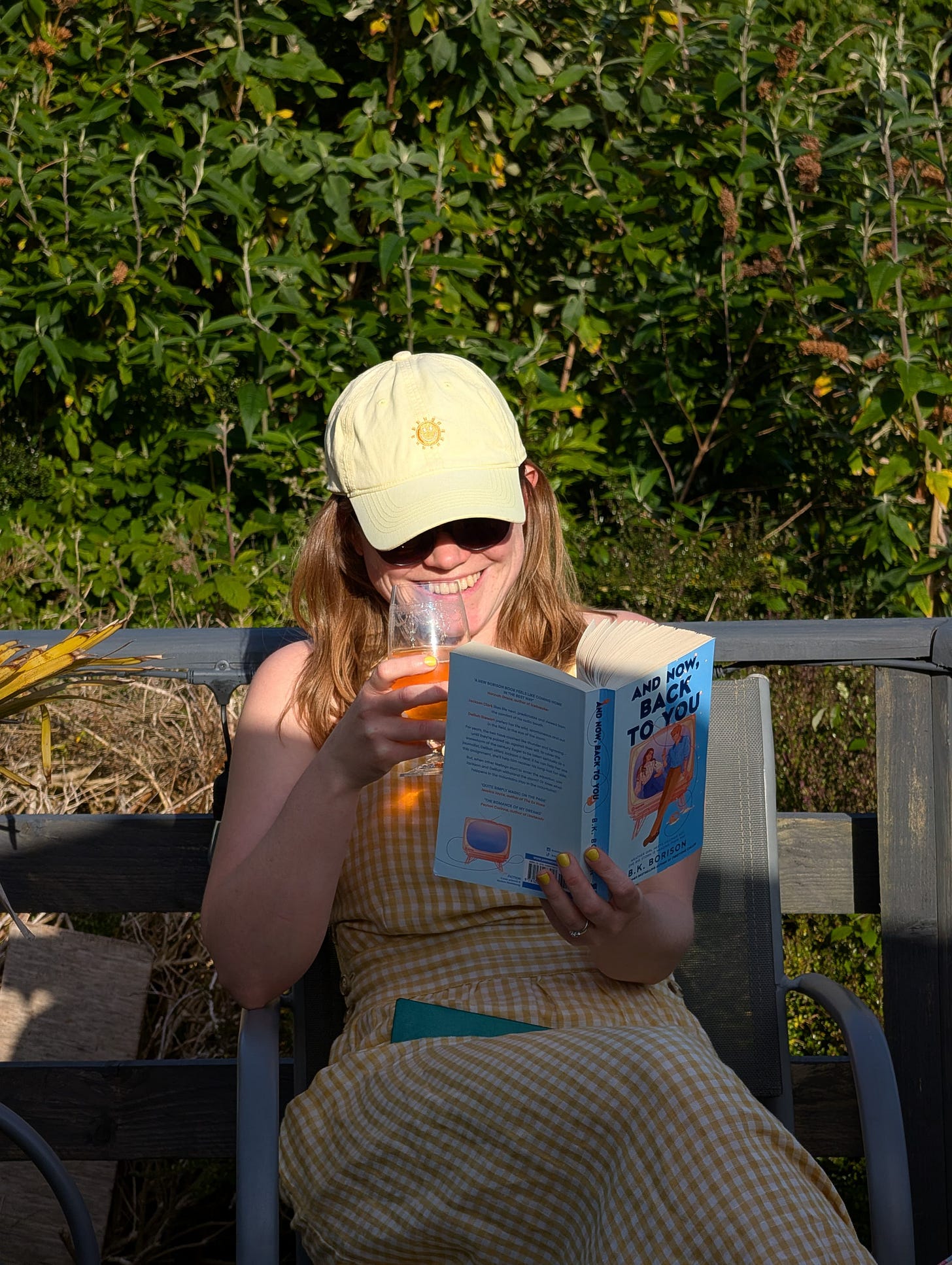 Libby, a white woman wearing a yellow gingham dress and yellow cap, sits in a chair reading, holding a drink and smiling Libby, a white woman wearing a yellow gingham dress and yellow cap, sits in a chair reading, holding a drink and smiling
