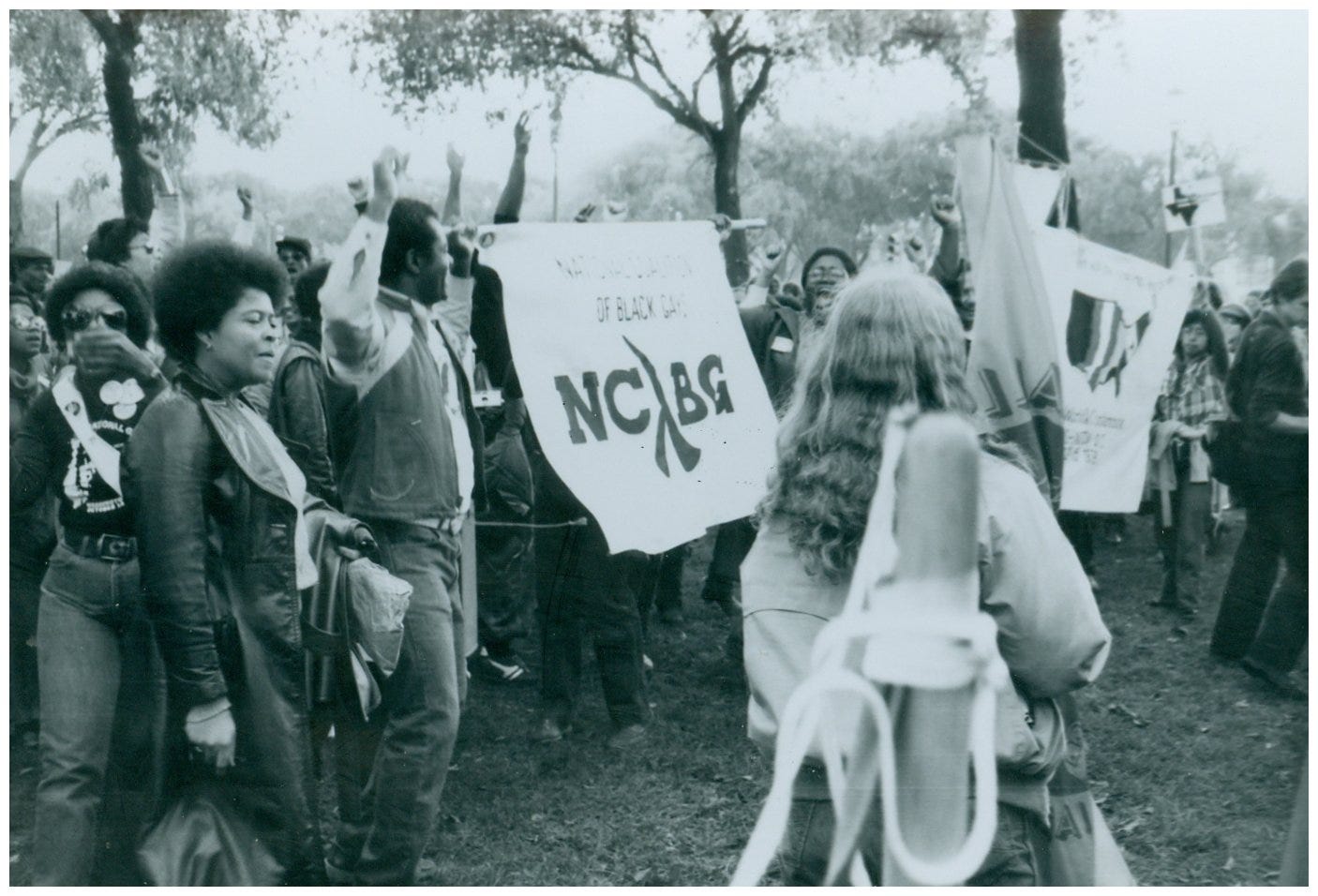 Black-and-white photograph of marchers gathered on the National Mall during the 1979 National March on Washington for Lesbian and Gay Rights. Members of the National Coalition of Black Gays stand with raised fists beside a banner reading “NCBG,” while another banner from the Third World Lesbian and Gay Conference is visible in the background. Participants appear joyful and resolute amid a crowd of demonstrators.