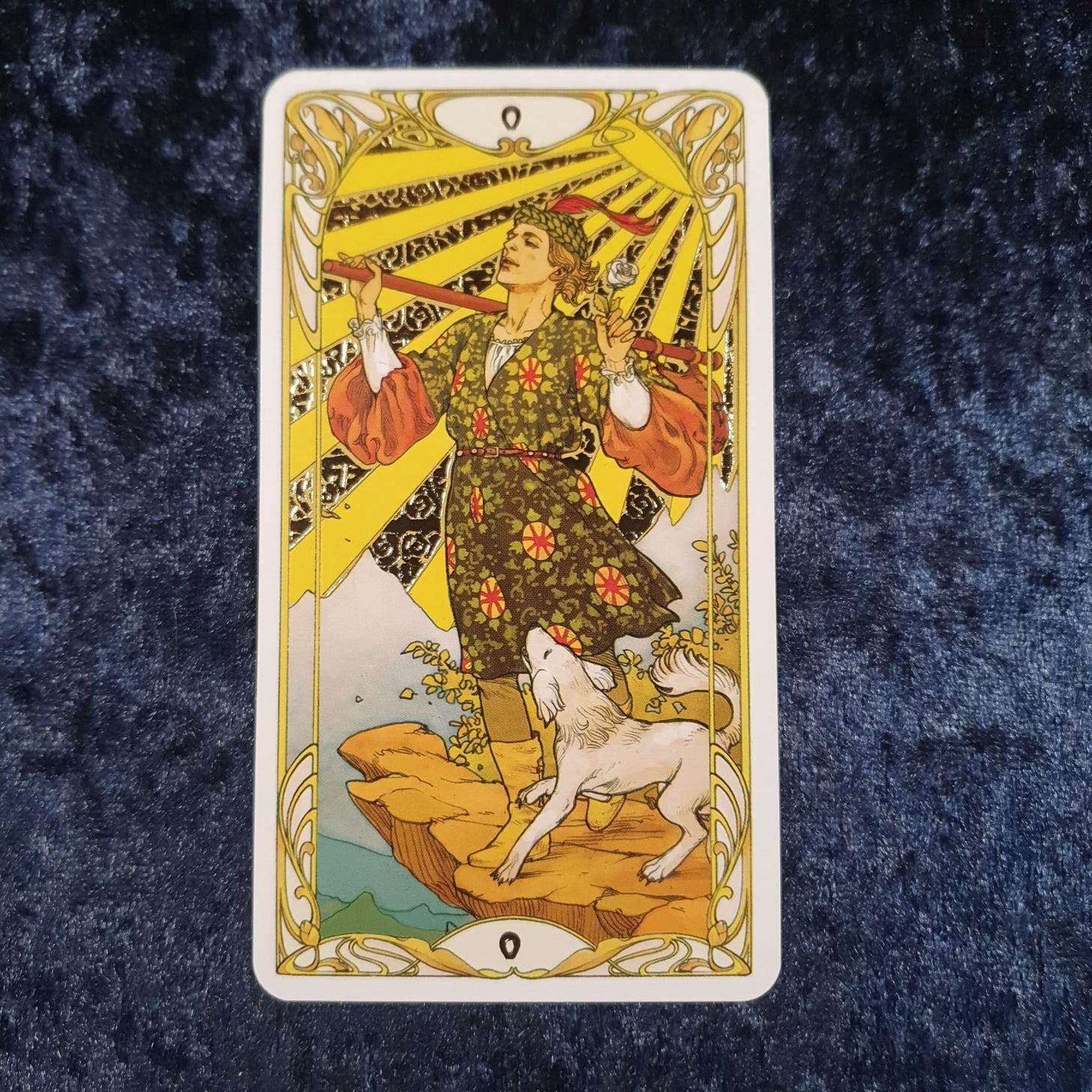 Alt text: The Fool from the Golden Art Nouveau Tarot on deep blue velvet, featuring a bright sunburst, white dog, and decorative gold linework, photographed for Tarot DMs with novelist Emma van Straaten.