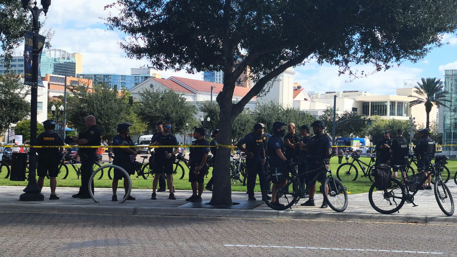 A group of police officers and bicycles gathered behind yellow caution tape in a park area.
