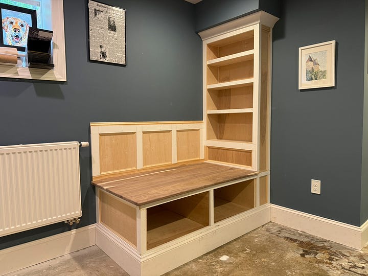A four-photo collage showing the later stages of building a custom reading nook and bookshelf. The top-left image shows a built-in bench with a walnut top and an adjacent floor-to-ceiling bookshelf installed beneath a ceiling bulkhead. The top-right image features the partially completed bench with a cat lounging on the seat and a black dog standing in the foreground. The bottom-left image shows additional trim and paneling added to the bench back and bookshelf, with the project taking shape within the blue-walled room. The bottom-right image shows further progress on the built-in, including decorative trim, shelving, and storage cubbies beneath the bench. Together, the photos document the transformation from basic cabinetry to a finished custom reading nook.