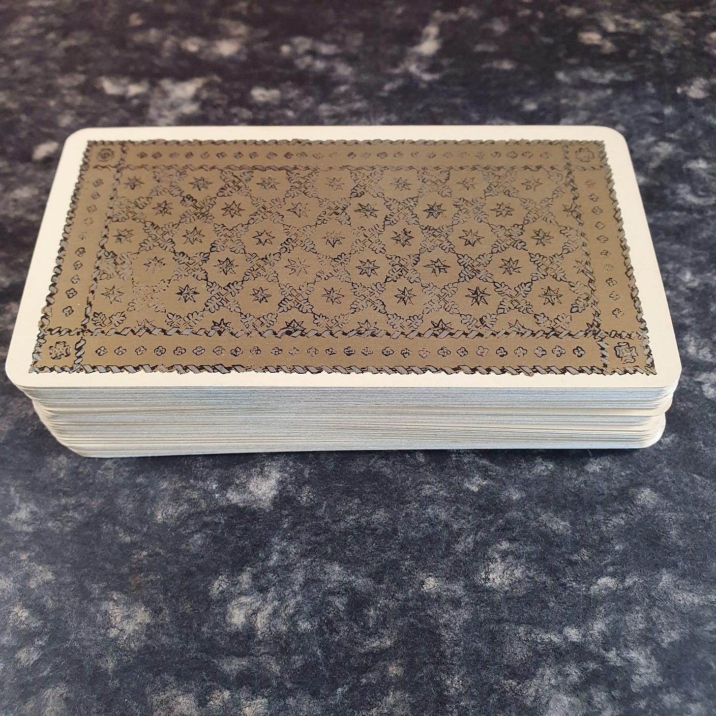 A thick tarot deck rests face down on a dark mottled surface, its card backs decorated with a brown lattice and star pattern. A thick tarot deck rests face down on a dark mottled surface, its card backs decorated with a brown lattice and star pattern.