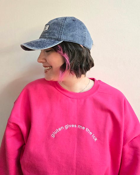 Modeling merch: A hot pink sweatshirt with white embroidered text on the front chest that says "gluten gives me the ick" and "GFGC" on the left sleeve near the wrist. A vintage wash denim baseball cap with an embroidered white pixelated chat bubble and black "GFGC" inside. A black sweatshirt with a green embroidered design on the front chest that says "GAME OVER, YOU ATE GLUTEN AND DIED" with a pixelated ox and wagon and "GFGC" on the left sleeve near the wrist.