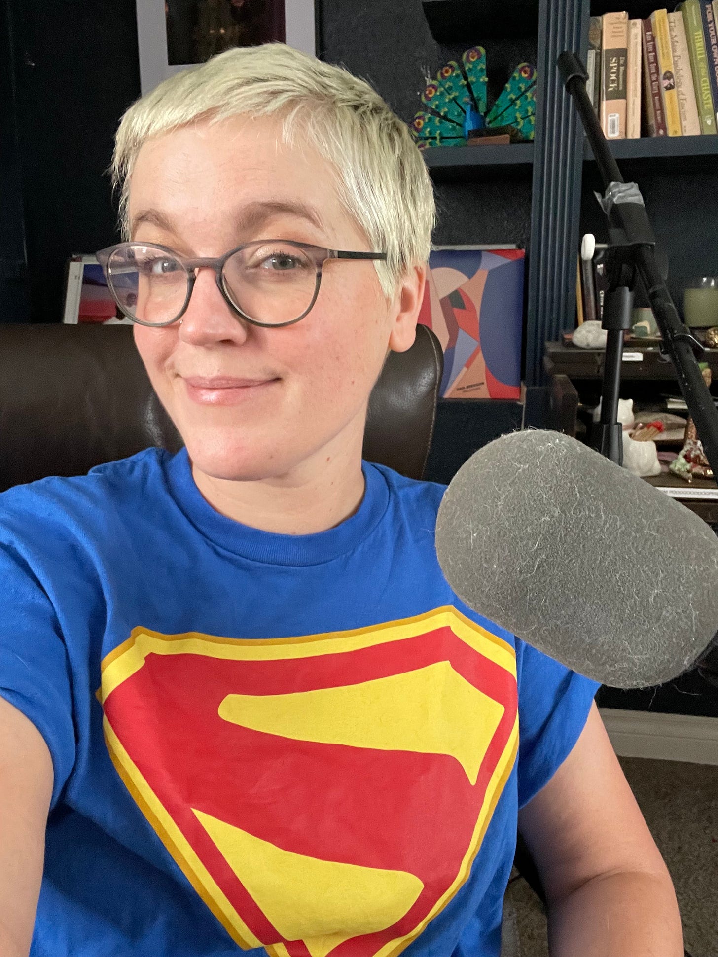 a white person with short bleached hair and glasses is smirking at the camera behind a microphone. they are wearing a superman tshirt