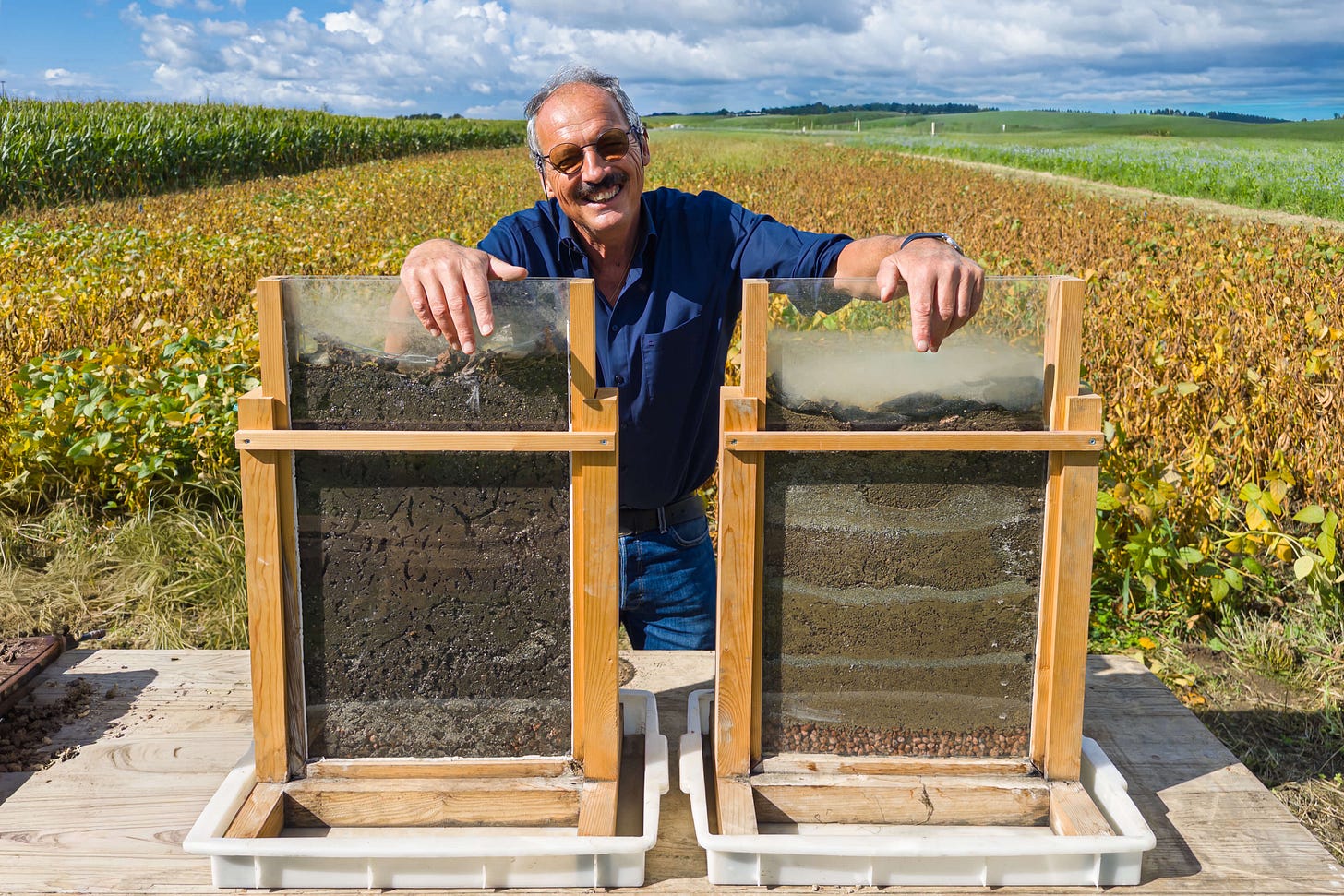 Andreas Chervet demonstriert auf dem Bodenacker bei Zollikofen einen Regenwurm-Bodentest zur Veranschaulichung der Bodenqualität. Er steht hinter zwei durchsichtigen Glasrahmen mit Bodensäulen: links ein dunkler, stark durchwurzelter und von Regenwürmern durchzogener Boden, rechts eine deutlich geschichtete, kompaktere Bodenprobe. Im Hintergrund sind Felder mit Sojabohnen und Mais zu sehen, darüber ein blauer Himmel mit Wolken.