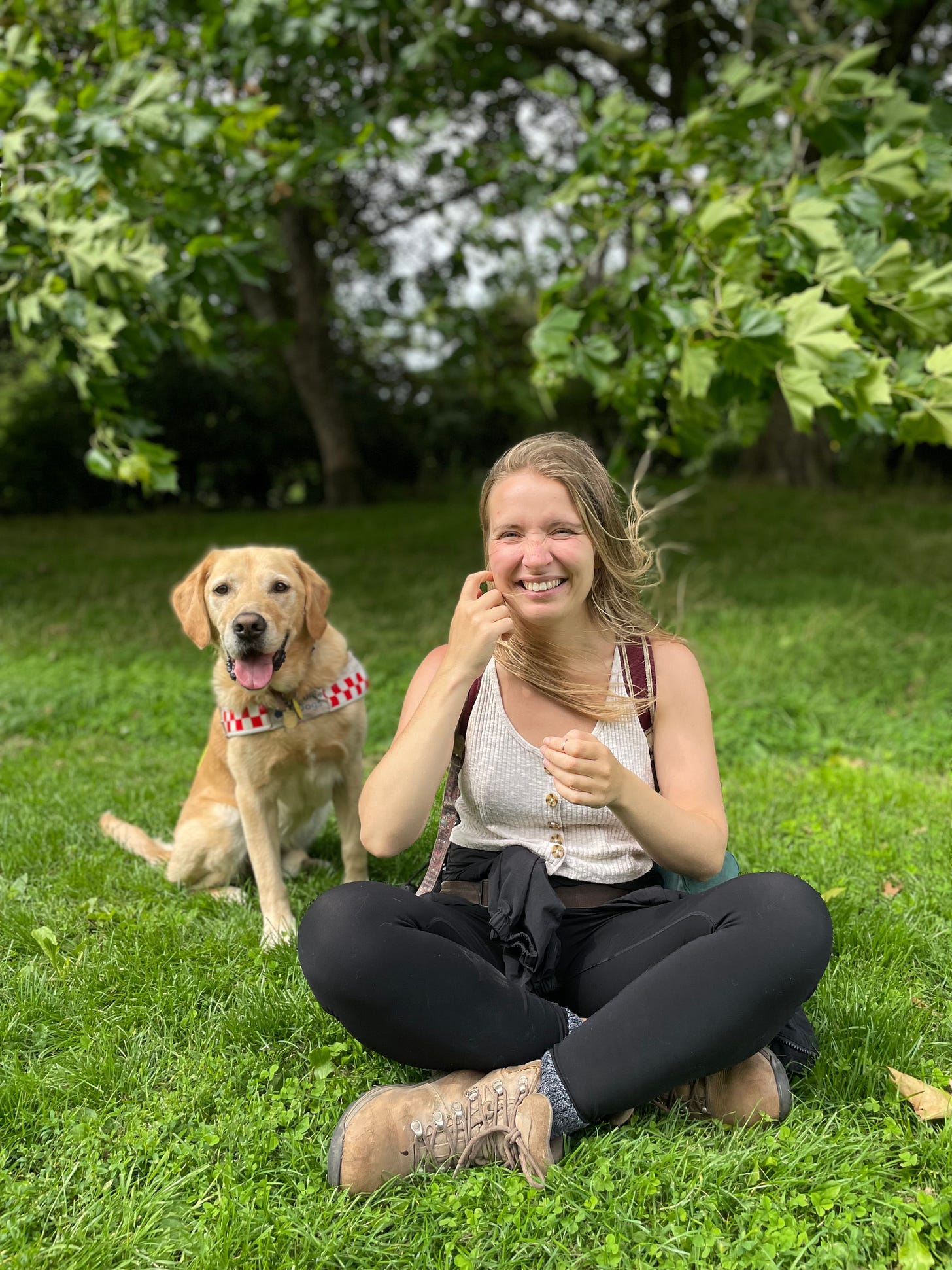 A similar photo of me and my guide dog sitting on a grass, but I am looking and smiling at the camera instead.