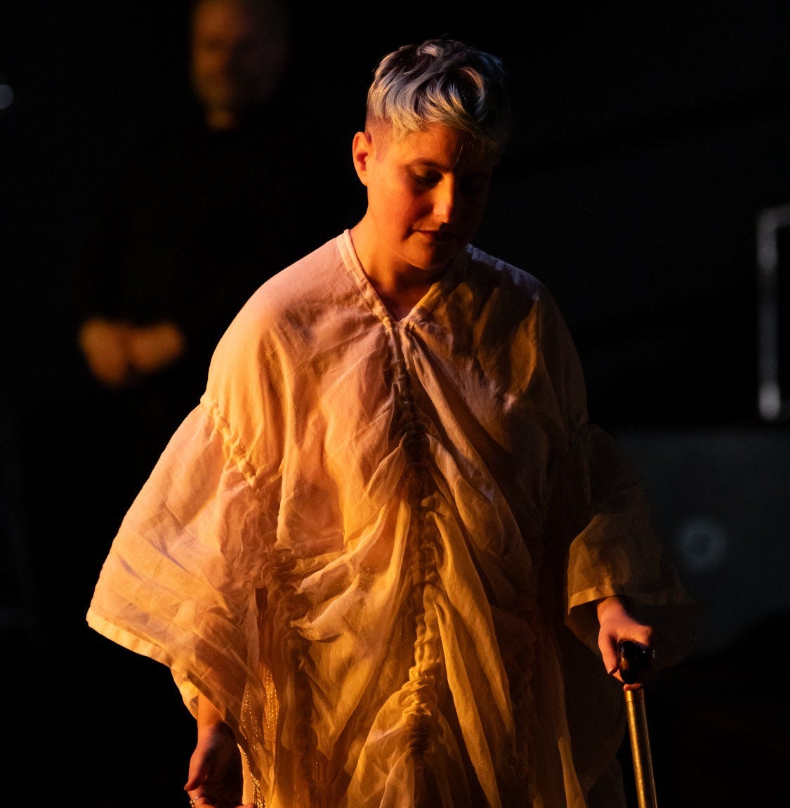 A photo of a white queer Jewish person wearing a long white robe or dress, using a cane in the warm glow of stage lights.