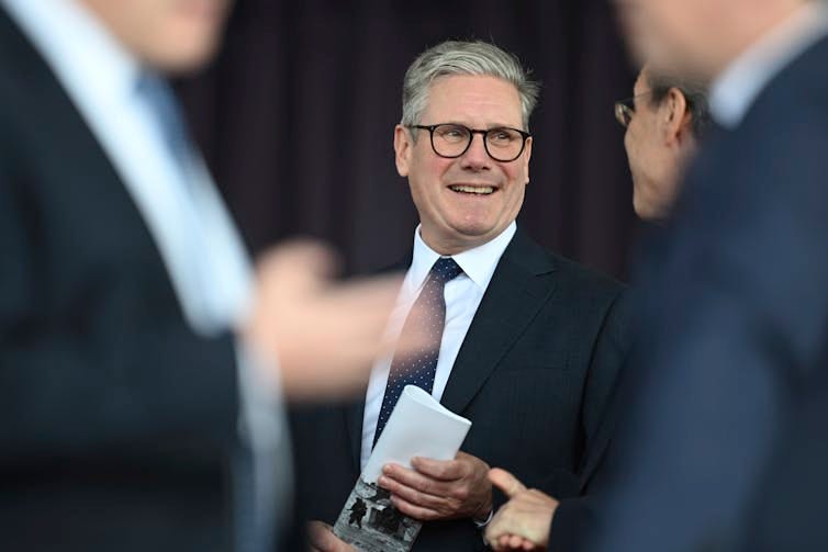 A man wearing glasses and in a dark suit and tie smiles with two blurred men in the foreground standing in front of him. A man wearing glasses and in a dark suit and tie smiles with two blurred men in the foreground standing in front of him.