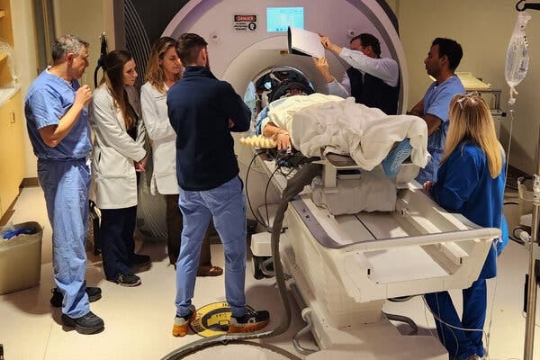 A patient in a hospital gown and blanket lies horizontal on an M.R.I. machine while several doctors and scientists crowd around during an experiment.