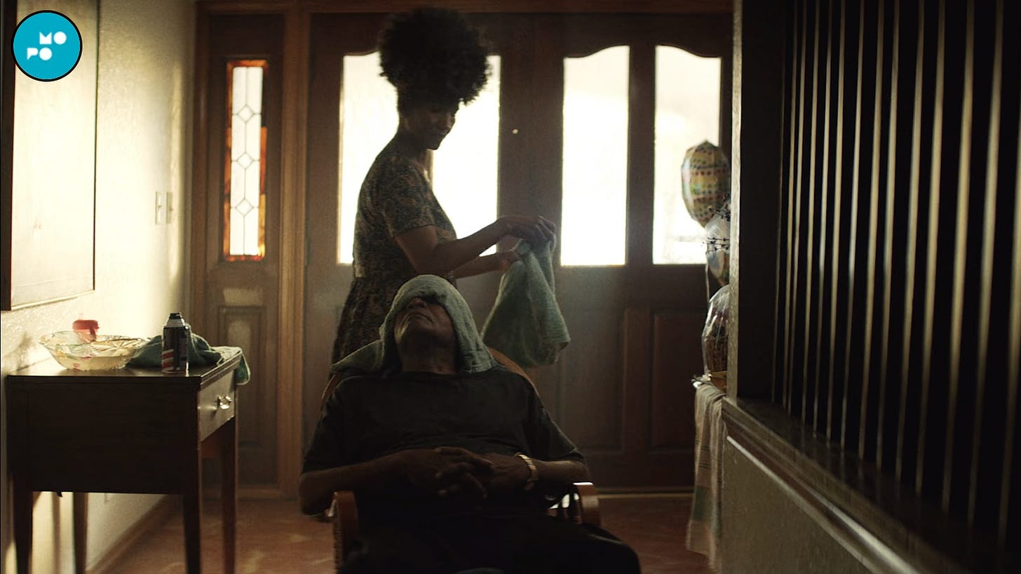 A scene from a film; two figures stand in a low-lit entryway, one is shaving the other. 